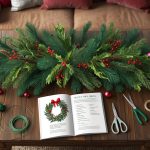 A cozy living room with a large wooden table covered in pine branches, holly berries, and assorted ribbons and ornaments. A wreath-making guidebook is open next to a pair of scissors and floral wire